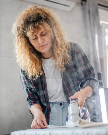 Téléchargez les photos : Femme adulte caucasienne heureuse femme utilisant du fer électrique pour repasser les vêtements à la maison tâches ménagères concept de vraies personnes travail domestique copie espace - en image libre de droit