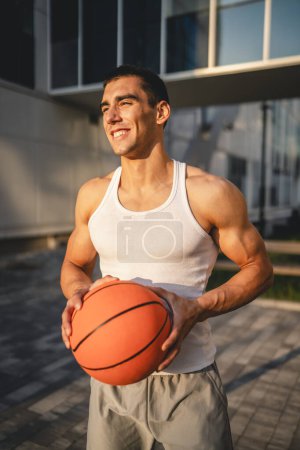 Téléchargez les photos : Un jeune homme caucasien athlète masculin debout extérieur tenir ballon de basket-ball porter blanc débardeur un-shirt forte musculaire personne réelle copie espace heureux sourire confiant mode de vie sain concept - en image libre de droit
