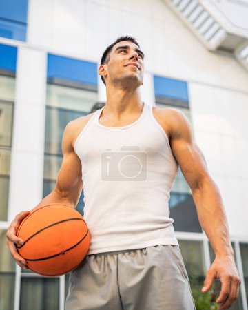 Téléchargez les photos : Un jeune homme caucasien athlète masculin debout extérieur tenir ballon de basket-ball porter blanc débardeur un-shirt forte musculaire personne réelle copie espace heureux sourire confiant mode de vie sain concept - en image libre de droit