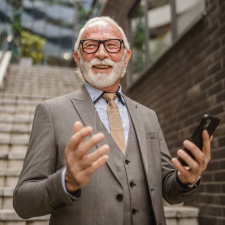 Téléchargez les photos : Un homme d'affaires senior debout en plein air dans un bâtiment moderne utiliser téléphone mobile smartphone pour passer un appel ou envoyer des SMS SMS textos personnes réelles costume de vêtements de cheveux gris - en image libre de droit