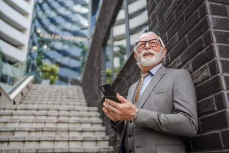 Téléchargez les photos : Un homme d'affaires senior debout en plein air dans un bâtiment moderne utiliser téléphone mobile smartphone pour passer un appel ou envoyer des SMS SMS textos personnes réelles costume de vêtements de cheveux gris - en image libre de droit