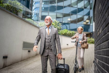 Téléchargez les photos : Senior couple homme et femme en face de l'hôtel bâtiment moderne avec valise et coup de pied scooter voyage d'affaires ou vacances concept homme d'affaires et femmes d'affaires amis ou mari et femme - en image libre de droit