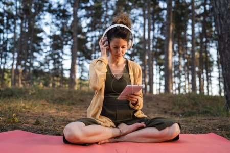 Foto de Una mujer joven adulta caucásica sentada en la esterilla de yoga sola en el parque o bosque en la naturaleza sosteniendo la tableta digital y ajustando los auriculares preparándose para la meditación guiada en línea autocuidado - Imagen libre de derechos