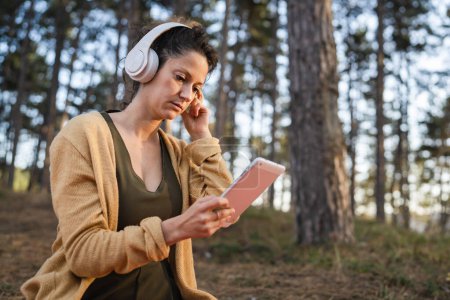 Photo for One woman young adult caucasian female sitting alone in the park or forest in nature holding digital tablet and adjusting headphones preparing for online guided meditation self-care practices concept - Royalty Free Image