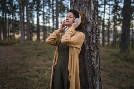 Foto de Una mujer joven adulta caucásica sentada sola en el parque o bosque en la naturaleza ajusta los auriculares preparándose para la meditación guiada auto-cuidado manifestación práctica equilibrio emocional mental concepto - Imagen libre de derechos