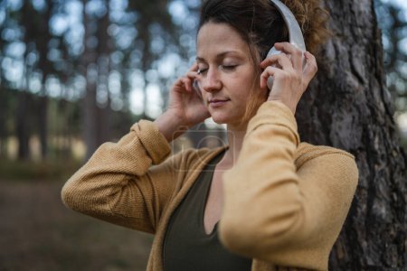 Photo for One woman young adult caucasian female sitting alone in the park or forest in nature adjust headphones preparing for guided meditation self-care manifestation practice mental emotional balance concept - Royalty Free Image