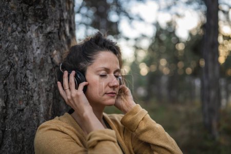 Téléchargez les photos : Une femme jeune femme caucasienne adulte assise seule dans le parc ou dans la forêt dans la nature avec écouteurs se préparant à la méditation guidée autosoin pratique concept d'équilibre émotionnel mental - en image libre de droit