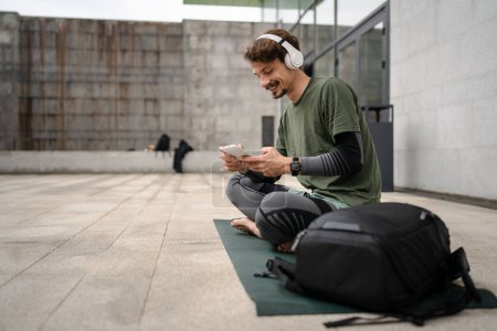 Photo for One man young adult caucasian male prepare for guided training yoga or meditation hold digital tablet while sitting outdoor with headphones self-care practice well-being inner peace balance concept - Royalty Free Image