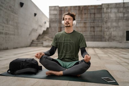 Téléchargez les photos : Un homme jeune adulte caucasien yeux masculins fermés pour la formation guidée yoga ou méditation tout en étant assis en plein air avec casque pratique d'auto-soins de vraies personnes bien-être concept de paix intérieure et d'équilibre - en image libre de droit