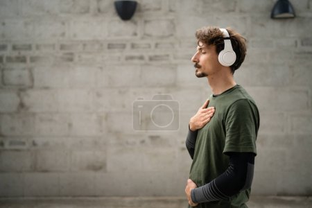 Téléchargez les photos : Un homme jeune adulte caucasien mâle vue de côté portrait de la personne moderne avec des moustaches porter des écouteurs regardant vers la caméra personnes réelles copier l'espace tour de taille debout concept de méditation guidée en plein air - en image libre de droit