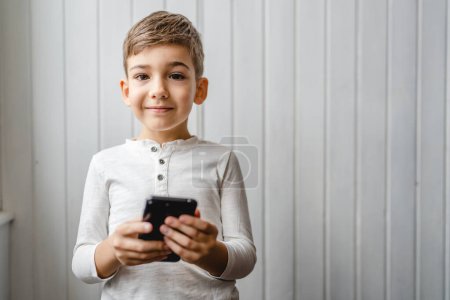 Foto de One boy caucasian child preschooler hold smartphone mobile phone at home play video games childhood and growing up technology addiction concept use smartphone app for online browsing or watch video - Imagen libre de derechos