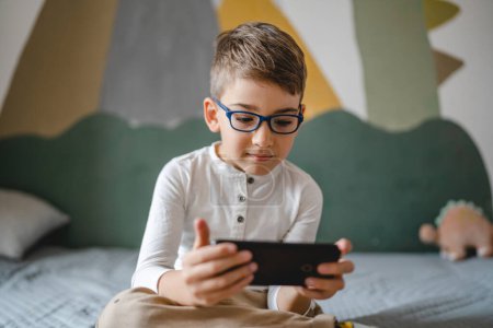 Téléchargez les photos : Un garçon caucasien enfant d'âge préscolaire tenir smartphone téléphone mobile à la maison jouer à des jeux vidéo enfance et grandir concept de dépendance à la technologie utiliser l'application smartphone pour la navigation en ligne ou regarder la vidéo - en image libre de droit