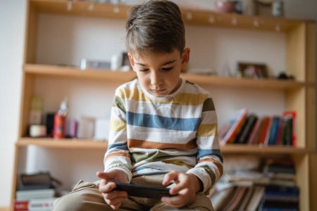 Téléchargez les photos : One boy caucasian child preschooler hold smartphone mobile phone at home play video games childhood and growing up technology addiction concept use smartphone app for online browsing or watch video - en image libre de droit