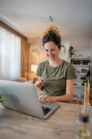 Téléchargez les photos : Une femme adulte caucasienne assise à la maison sourire heureux à l'ordinateur portable tenir carte de crédit bancaire concept de magasinage en ligne personnes réelles copier l'espace e-commerce - en image libre de droit