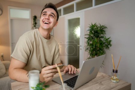 Foto de One man adult caucasian male sit at home happy smile at laptop computer hold bank credit card online shopping concept real people copy space e-commerce - Imagen libre de derechos