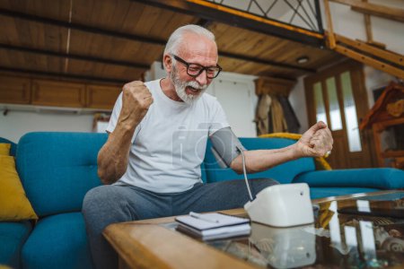 Photo for One man happy senior caucasian male with beard healthy celebrate use blood pressure device to check and measure results while sit at the table at home alone real people copy space health care concept - Royalty Free Image