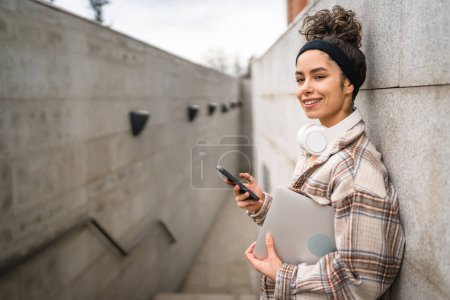 Téléchargez les photos : Une femme jeune adulte étudiante debout ou marcher en plein air tenir ordinateur portable utiliser téléphone intelligent pour SMS messages texte en ligne personne réelle copier l'espace heureux sourire confiant avec écouteurs dans la journée - en image libre de droit