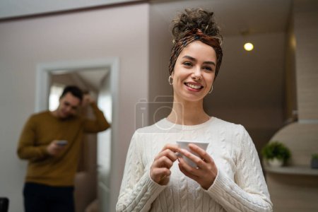 Téléchargez les photos : One woman young caucasian female standing in her apartment with cup of coffee while her husband or boyfriend stand in background daily morning evening couple routine real people copy space happy smile - en image libre de droit