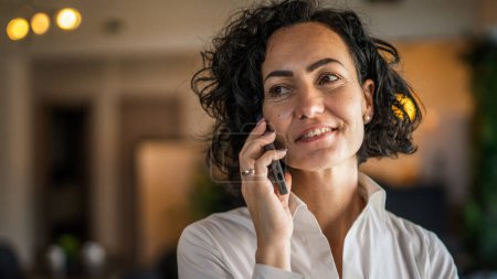 Foto de One woman mature caucasian female businesswoman entrepreneur stand at work or home use mobile phone making a call talk real people copy space wear white shirt curly hair happy smile - Imagen libre de derechos