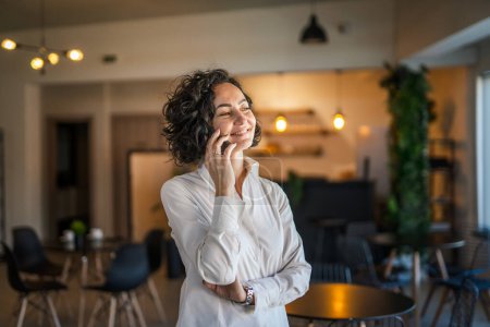 Foto de One woman mature caucasian female businesswoman entrepreneur stand at work or home use mobile phone making a call talk real people copy space wear white shirt curly hair happy smile - Imagen libre de derechos