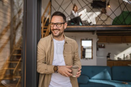 Téléchargez les photos : Un homme adulte caucasien mâle avec barbe et lunettes debout sur le balcon à la porte dans la journée heureux sourire tenir tasse de café copie espace matin routine quotidienne concept - en image libre de droit