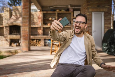 Téléchargez les photos : Un homme heureux caucasien masculin utiliser smartphone pour selfie photo ou vidéo appel extérieur en face de la maison minuscule moderne tout en vacances dans la journée les gens réels copier l'espace - en image libre de droit