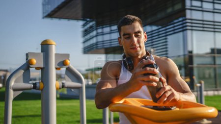 Téléchargez les photos : Un homme caucasien jeune athlète masculin prendre un frein pendant l'entraînement en plein air dans le parc salle de gym en plein air tenir shaker supplément dans la main heureux confiant espace de copie forte - en image libre de droit