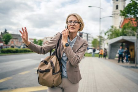 Photo for One woman caucasian mature blonde female with eyeglasses stand at bus city stop in day use smartphone mobile phone making a call talk while wait for the drive real person copy space late for work - Royalty Free Image