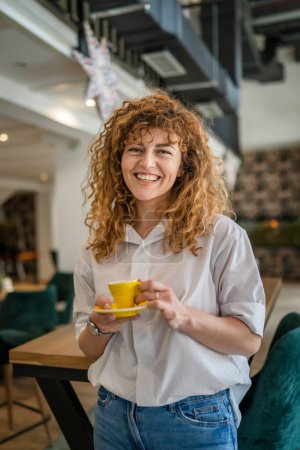 Foto de Una mujer caucásica adulta con sonrisa de pelo rizado tomando una taza de café en la cafetería sin preocupaciones disfrutar de espacio de copia sonriente feliz - Imagen libre de derechos