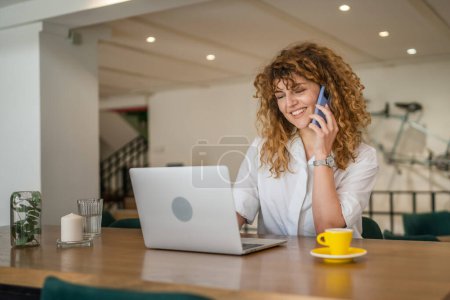 Foto de Una mujer adulta caucásica hembra sentarse en el trabajo de la cafetería en el ordenador portátil hacer una llamada telefónica hablar sonrisa feliz confiado sonriendo espacio de copia en persona real - Imagen libre de derechos