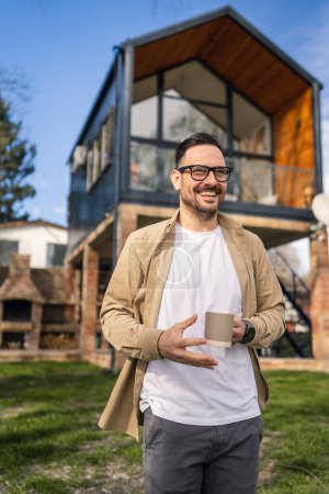 Photo for A 40-year-old adult man stands confidently in front of his modern home holding a cup of tea or coffee smiling He looks content and happy with his life and proud of his house copy space - Royalty Free Image