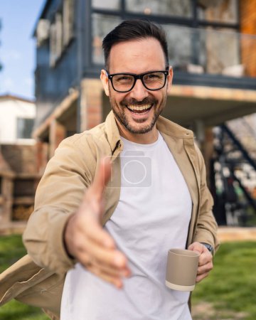 Photo for A 40-year-old adult man stands confidently in front of his modern home holding a cup of tea or coffee smiling He looks content and happy with his life and proud of his house copy space - Royalty Free Image