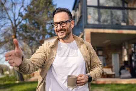 Téléchargez les photos : Un homme adulte de 40 ans se tient en confiance devant sa maison moderne tenant une tasse de thé ou de café souriant Il a l'air content et heureux de sa vie et fier de son espace de copie de maison - en image libre de droit