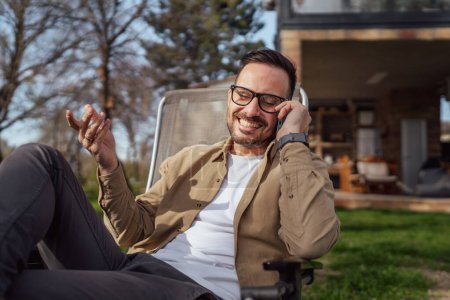 Photo for One adult man 40 years old caucasian male sit in front of his house on vacation talk on his smartphone mobile phone call happy smile during the conversation real person copy space - Royalty Free Image