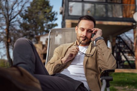 Photo for One adult man 40 years old caucasian male sit in front of his house on vacation talk on his smartphone mobile phone call during the conversation real person copy space - Royalty Free Image