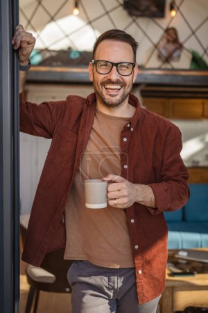 Photo for Man caucasian 40 years old male wear a shirt stands on a balcony holding a cup of tea or coffee smiling happy He is enjoying a peaceful moment in his daily morning routine or on a vacation copy space - Royalty Free Image