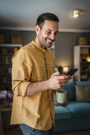 Foto de Un hombre adulto caucásico uso masculino teléfono móvil teléfono inteligente para enviar mensajes de texto SMS o navegar por Internet, mientras que de pie en casa llevar camisa y gafas persona real copiar espacio sonrisa feliz - Imagen libre de derechos