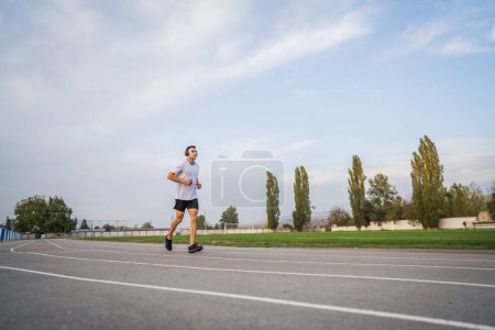 Téléchargez les photos : Adulte homme caucasien jogging sur la piste de course athlète masculin dans l'entraînement du stade courir au printemps ensoleillé ou jour d'été de vraies personnes concept de mode de vie sain - en image libre de droit