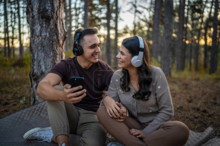 Téléchargez les photos : Homme et femme jeune couple adulte dans la nature écouter de la musique sur casque utiliser un téléphone portable smartphone ou faire une application d'appel vidéo - en image libre de droit