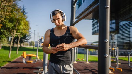 Photo for One man caucasian young male stand at outdoor open training park gym use mobile phone smartphone with headphones send messages texting or browse internet online app for training real person copy space - Royalty Free Image