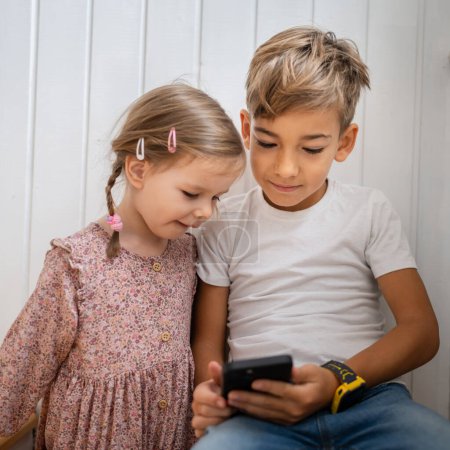 Photo for Siblings brother boy hold smartphone with girl sister children use mobile phone smartphone at home in room watch video make a call or play online games leisure family concept real people copy space - Royalty Free Image