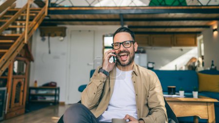 Téléchargez les photos : Un homme caucasien mâle avec barbe et lunettes assis à la maison utiliser téléphone intelligent mobile tandis que le travail de l'espace de copie à la maison les gens réels parlent - en image libre de droit