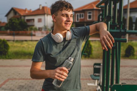 Téléchargez les photos : Un homme caucasien adolescent masculin tenir bouteille en plastique d'ouverture d'eau et se préparer à boire tout en s'entraînant en plein air dans l'hydratation du jour ensoleillé et un mode de vie sain concept copier l'espace - en image libre de droit