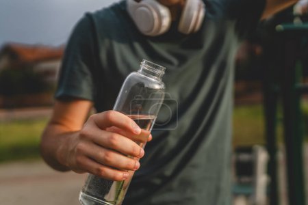 Téléchargez les photos : Un homme caucasien adolescent masculin tenir bouteille en plastique d'ouverture d'eau et se préparer à boire tout en s'entraînant en plein air dans l'hydratation du jour ensoleillé et un mode de vie sain concept copier l'espace - en image libre de droit