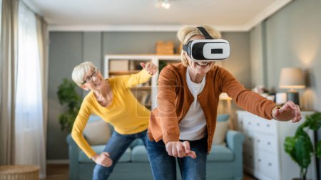 Foto de Dos mujeres maduras mayores caucásicas amigas o hermanas en casa disfrutar de realidad virtual VR auriculares personas reales activos senior divertirse concepto de ocio - Imagen libre de derechos