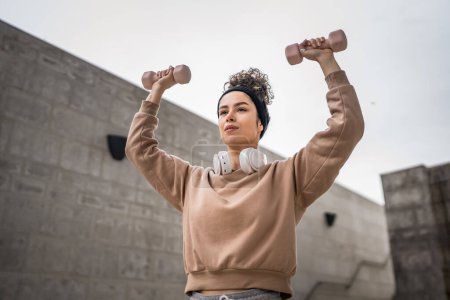 Téléchargez les photos : Femme caucasienne adulte s'entraînant en plein air dans la ville jour athlète féminine en utilisant des haltères poids dans sa routine d'entraînement quotidienne - en image libre de droit
