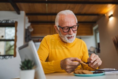 Photo for One senior man caucasian old male pensioner sit at a table at home putting jam on a biscuit preparing to eat breakfast enjoy snack relaxed in his home environment real person copy space daily routine - Royalty Free Image