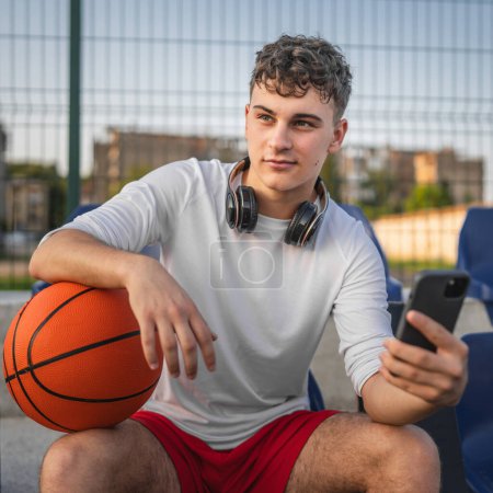 Téléchargez les photos : Caucasien adolescent en utilisant téléphone mobile smartphone à un terrain de basket - en image libre de droit