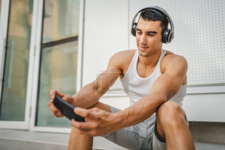 Téléchargez les photos : Portrait de jeune homme utilisation extérieure écouteurs et téléphone intelligent pour jouer à des jeux vidéo - en image libre de droit