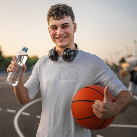 Téléchargez les photos : Homme caucasien adolescent ouvert bouteille en plastique d'eau en plein air dans une boisson ensoleillée jour tandis que le basket-ball trou - en image libre de droit